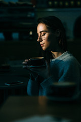 Close up erarly morning vertical portrait of a beautiful european woman drinking coffee with close eyes in a dark background. High contrast.
