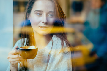 Young thoughtful woman sitting in a cafe and looking outside in winter time. Hipster female dressed in warm clothes.The photo was taken through the window.