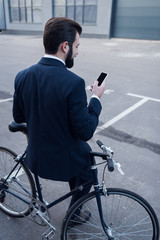back view of businessman using smartphone while leaning on bicycle on street
