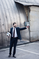 young smiling businessman in suit pointing away while playing soccer on street