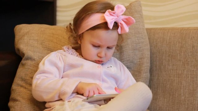 Funny child looks at the phone screen and plays downloaded application on a smart phone close-up. A little cute girl lies in sofa in a living room, looking cartoon and playing the game