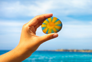 Person hand hold pebble with painted bright sun against blue sky and sea.