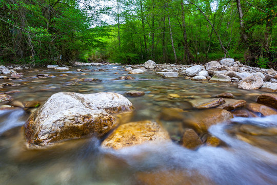 Transparent cold water of a mountain river flows between picturesque summer stones against a background of green trees close up. Forest landscape in the vicinity of Sochi, Russia.