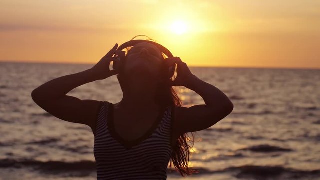 Young pretty woman wearing headphones have fun listening music on the beach by the sea at amazing sunset in slow motion. 1920x1080