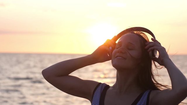 Young brunette woman wearing headphones have fun listening music on the beach at amazing sunset in slow motion. 1920x1080
