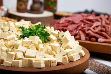 Close-up of big wooden plate, served with little cheese pieces and decorated with fresh parsley leaves. There is delicious sausage set on the background. Tasty appetizers for strong alcohol drinks.