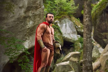 Portrait of a handsome Spartan warrior with strong muscular body wearing red cloak standing in the...