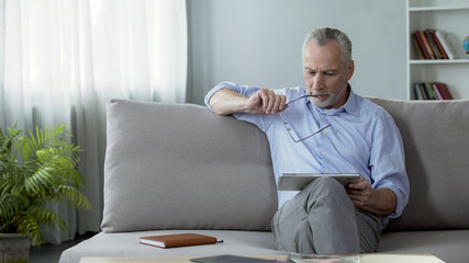 Adult male sitting on sofa and reading news on tablet, modern technologies