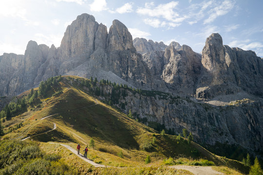 Two hikers walking into the mountains along a small trail with a beautiful panorama of high rocky mountain peaks and green fields and trees ahead of them