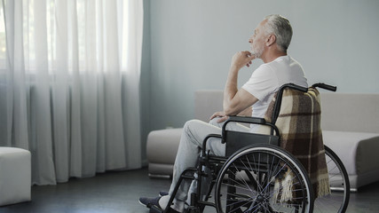 Thoughtful retired male sitting in wheelchair and looking in the window, disease