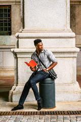 African American College Student studying on street in New York, wearing gray shirt, jeans, cloth shoes, wristwatch, carrying shoulder leather bag, listening music with earphone, reading red book..