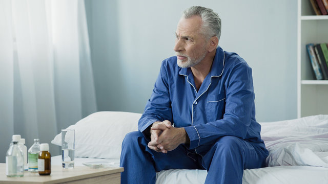 Serious aged man sitting upset and pensive on bed at home, lonely sick person