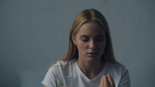 Caring about health. Close up of young girl holding a glass of water and looking at it while hesitating before taking a pill