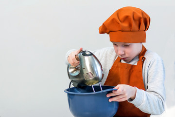 The blond boy in the orange dress of the chef stirs something with a mixer in a blue cup on a light background.