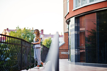 Portrait of a perfect young woman wearing striped overall and yellow sunglasses poses with her handbag on a balcony of a modern building in a town.