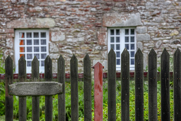 A pretty stone cottage with stone lintels and white mullioned windows stands surrounded by grass and a picket fence.