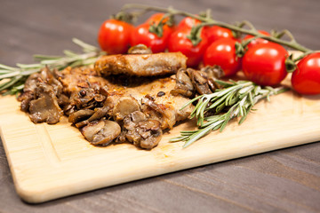 Close up on wooden board with grilled mushrooms and pork steak next to cherry tomatoes on a wooden table