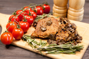 Delicious pork steak on wooden board next to a branch of oregano and cherry tomatoes. Two shakers with salt and pepper are blurred in the background