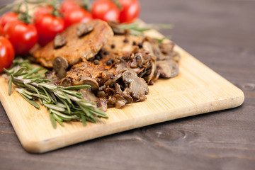 Tasty pork meat with grilled mushrooms next to oregano branch on wooden board. Cherry tomatoes are in blurred backgorund