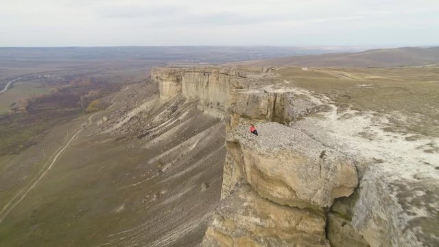 Young woman is sitting on the edge of tall wall of rock. Aerial view. Drone is flying forward over model. Establishing shot.