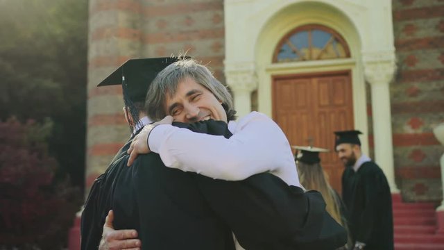 Young man hugging with his happy dad when he congratulating his son after the graduation ceremony. Outdoors