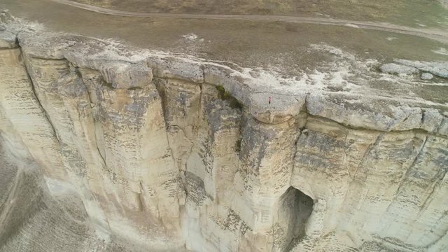 Woman is standing on the edge of huge tall wall of rock. Aerial view. Drone is flying backward and upward. Establishing revealing shot.