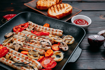 Grilled sausages and vegetables served in a grill pan with sauce and spices and crunchy wheat toasts on dark wooden table. Appetizing meat menu, kitchen background, restaurant concept