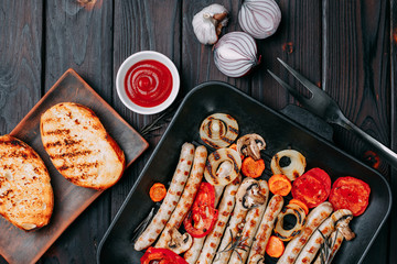 Grilled sausages and vegetables served in a grill pan with sauce and spices and crunchy wheat toasts, flat lay on dark wooden table. Appetizing meat menu, kitchen background, restaurant concept