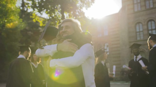 Rear of the blonde female graduate being congratulating with graduation by her gray-haired father on a sunny day. Outside