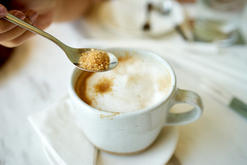 Woman's hand holding coffee spoon to pour brown sugar in a cup of coffee on white table