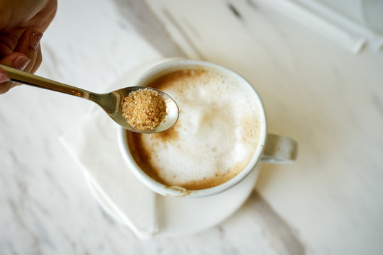 Woman's hand holding coffee spoon to pour brown sugar in a cup of coffee on white table