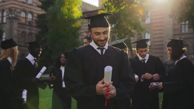 Portrait of the handsome man graduate standing in traditional graduation clothes with diploma in hands and looking to the sky with a hope. Graduates on the background. Outside