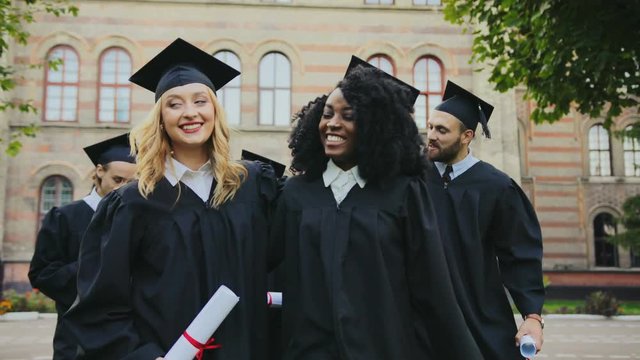 Happy smiled mixed races graduates walking with the diplomas in hands near the University building after the graduation ceremony. Outdoor
