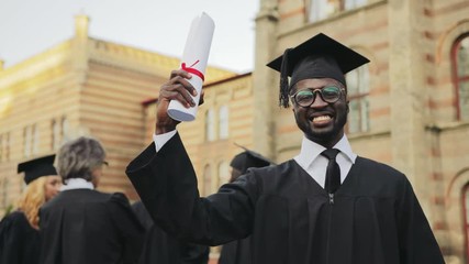 Portrait of the African American happy young graduated man posing to the camera and showing his diploma in front of the University. Graduates with professor on the background. Outdoors