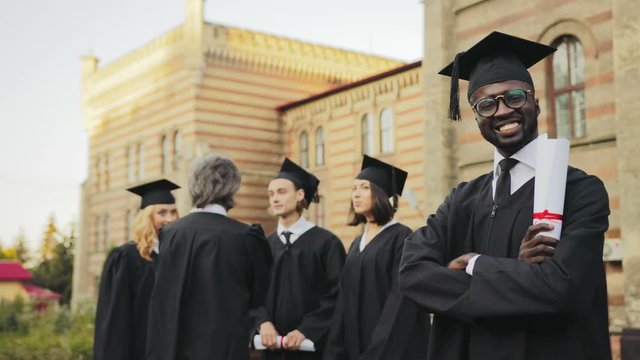 Portrait of the African American smiled male graduate posing to the camera and crossing his hands in front of the University. Graduates with professor on the background. Outdoor