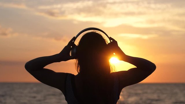 Silhouette of young woman wearing headphones have fun listening music on the beach at amazing sunset in slow motion. 1920x1080