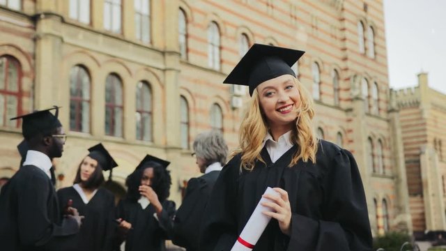 Portrait of the blonde young graduated woman posing to the camera and smiling in front of the University. Graduates with professor on the background. Outdoors