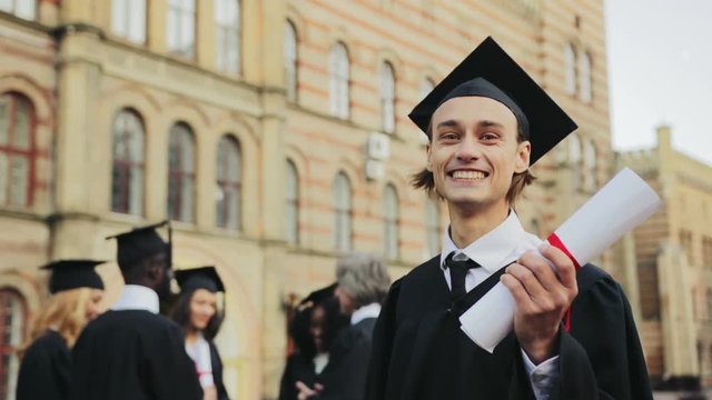 Portrait of the Caucasian happy young graduated man posing to the camera and showing his diploma in front of the University. Graduates with professor on the background. Outdoors