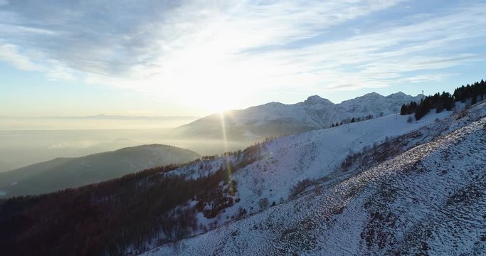 Forward aerial top view over winter snowy mountain and woods forest at sunset or sunrise.Dusk or dawn twilight sunshine flare.Mountains snow season sun establisher.4k drone flight establishing shot