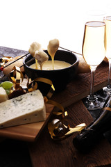 Gourmet Swiss fondue dinner on a winter evening with assorted cheeses on a board alongside a heated pot of cheese fondue with two forks dipping bread and white wine or champagne