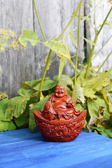 Ceramic buddha figurine on blue aged wooden boards in summer park with flowers.