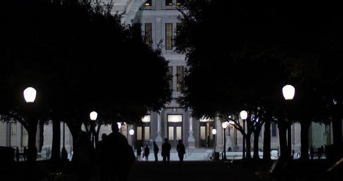 Silhouetted people walk in front of the Texas State Capitol Building in downtown Austin as night.  	