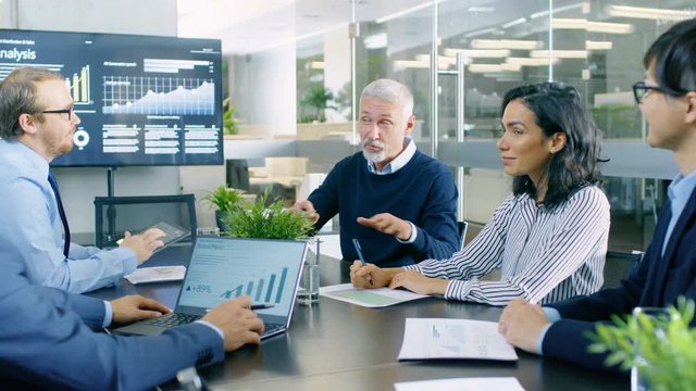 Senior Executive Explains Company's Vision and Potential to His Employees. They are Sitting at Big Table in Meeting Room. TV Screen on the Wall Shows Corporate Growth. RED EPIC-W 8K Camera.