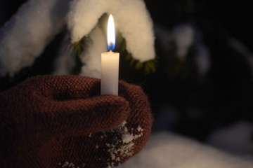 Obraz na płótnie Canvas Human hand with gloves hold a burning candle, with snow and tree in the dark background