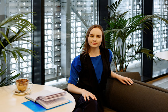 The Young Business Woman In A Blue Suit Sits At A Little Table In Cafe During A Business Lunch And Makes Entries In The Daily Log. The Cup Of Coffee Stands Nearby.