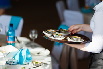 A waiter carries on a brown tray of food against the background of the decorated table. Hot snacks on the tray. The wait staff serving the tables at the event or holiday.