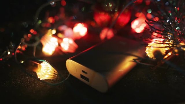 Silver powerbank external battery for fast charging smartphone or tablet pc lying on the table in the dark surrounded by flashing garland lights.