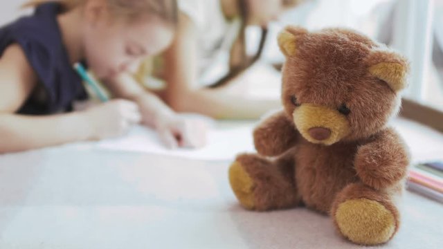 Teddy bear in close-up, two girls draws pictures at blurred background