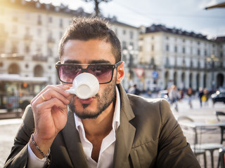 Handsome young man drinking espresso coffee, wearing elegant coat posing at table outside on urban...