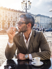 Handsome young man drinking espresso coffee and smoking cigarette, wearing elegant coat posing at...